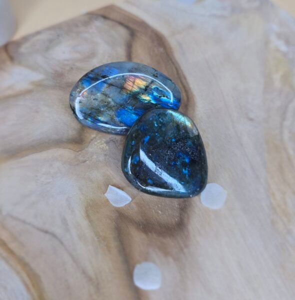 Deux pierres de labradorite polies aux reflets irisés, posées sur une planche en bois clair avec des pétales blancs autour.