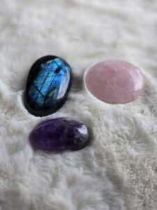 Trois pierres naturelles posées sur un tapis poils crème : Labradorite gris bleuté, Quartz rose tendre et Améthyste violette. Vue en flat lay, ambiance douce et épurée.
