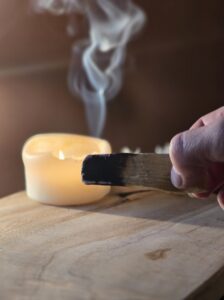 Main de Sophie, fondatrice d'Isha Kriya, tenant un bâton de Palo Santo fumant avec bougie allumée et petites fleurs blanches floutées, posés sur bois clair fond marron, rituel de purification énergétique
