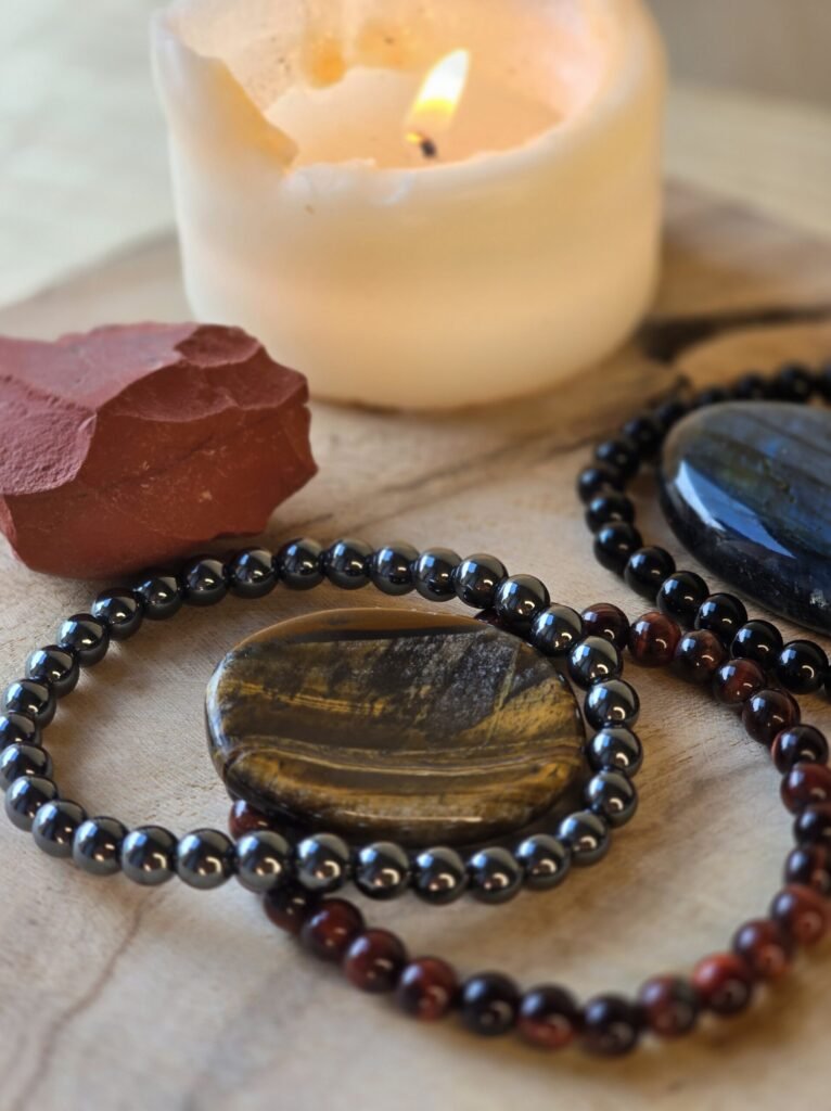 Ensemble de pierres naturelles pour protection énergétique et ancrage : tourmaline noire, obsidienne noire, labradorite, jaspe rouge, hématite, œil de tigre et œil de taureau, posées sur bois naturel avec bougie en arrière-plan