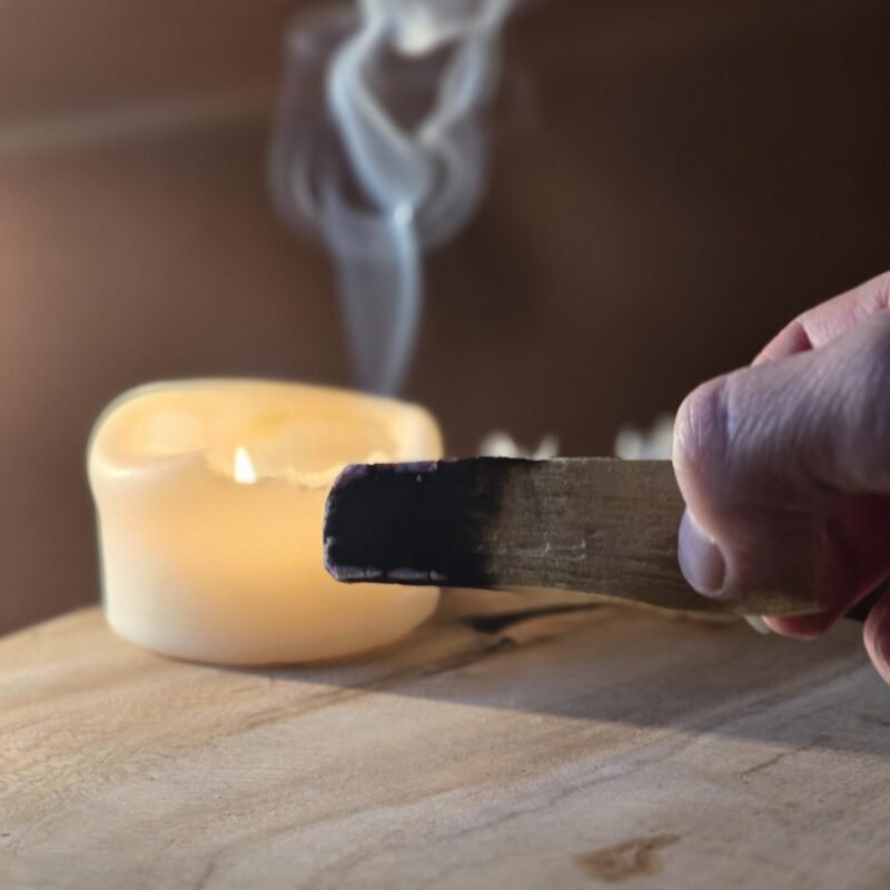 Main de Sophie, fondatrice d'Isha Kriya, tenant un bâton de Palo Santo fumant avec bougie allumée et petites fleurs blanches floutées, posés sur bois clair fond marron, rituel de purification énergétique