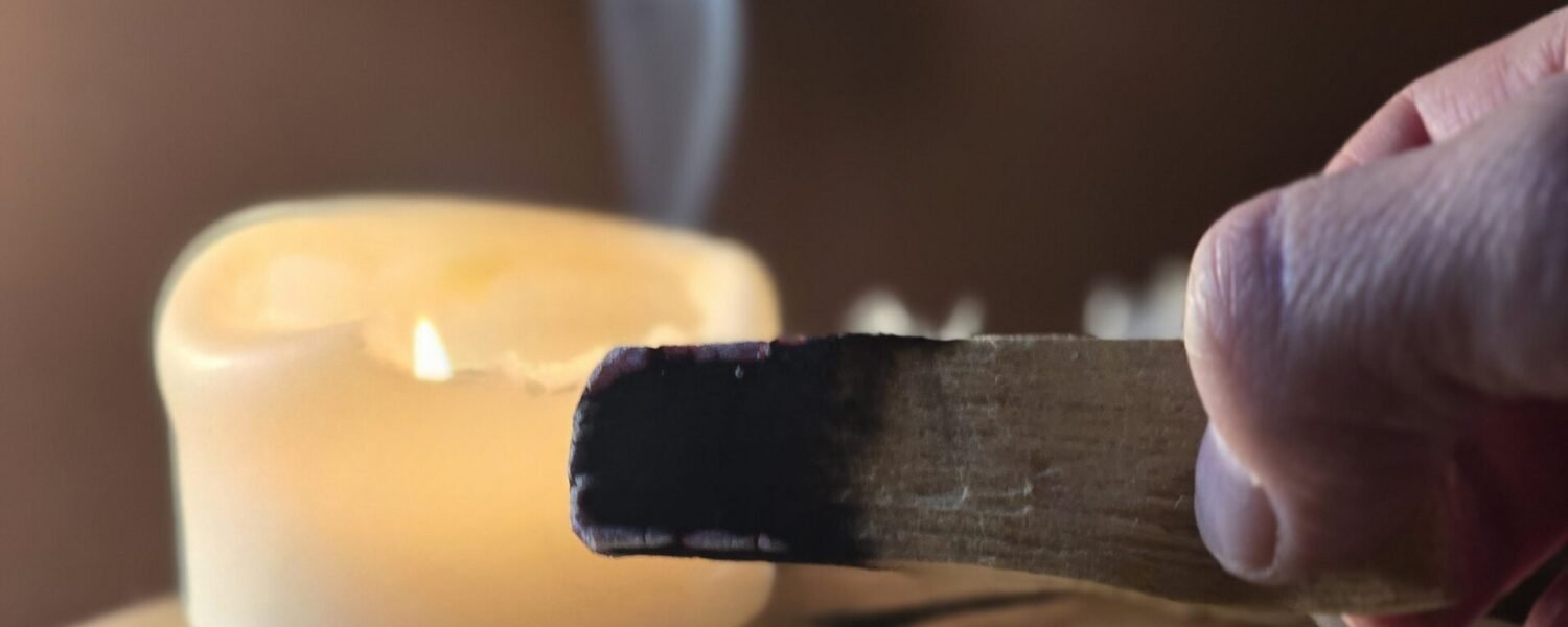 Main de Sophie, fondatrice d'Isha Kriya, tenant un bâton de Palo Santo fumant avec bougie allumée et petites fleurs blanches floutées, posés sur bois clair fond marron, rituel de purification énergétique