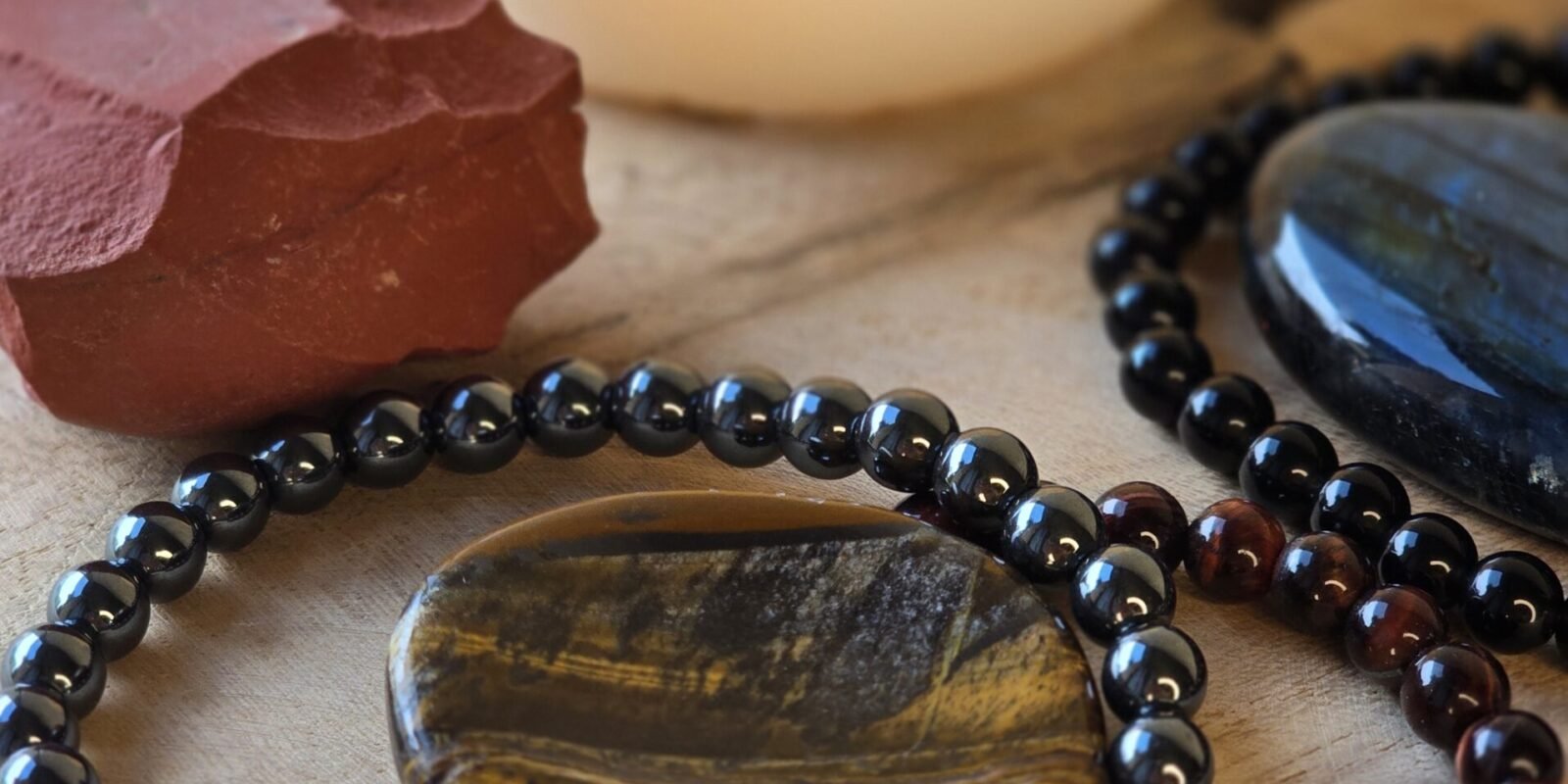 Ensemble de pierres naturelles pour protection énergétique et ancrage : tourmaline noire, obsidienne noire, labradorite, jaspe rouge, hématite, œil de tigre et œil de taureau, posées sur bois naturel avec bougie en arrière-plan